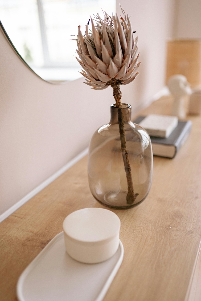 Auf einem Holzfach befinden sich ein weißes Tableau mit einer weißen Dose, eine bräunliche Glasvase mit Trockenblume und zwei aufeinanderliegende Bücher. Über dem Fach hängt ein runder Spiegel.