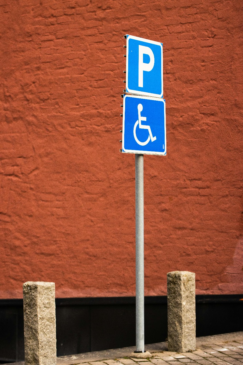 Schild zur Kennzeichnung eines barrierefreien Parkplatzes vor einer rot gefärbten Stein-Ziegel-Wand mit schwarzem Sockelbereich